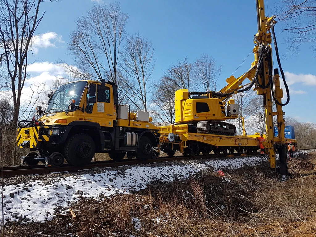 Gelbes Spezialfahrzeug führt Oberleitungsarbeiten in einem winterlichen Wald durch.