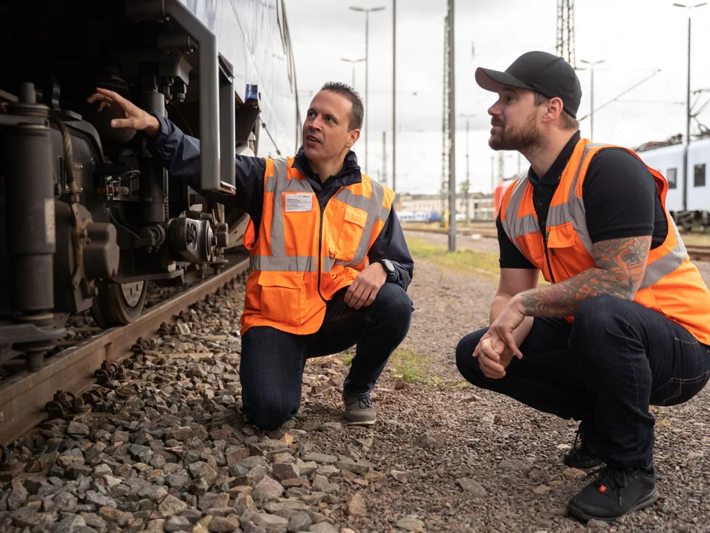 Mechatroniker und Kollegin in Warnkleidung prüfen Zugkomponenten auf einem Bahngelände.
